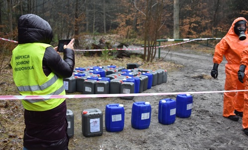 Inspektor Wojewódzkiego Inspektoratu Ochrony środowiska monitoruje pobór próbek z pojemników o pojemności 20 l porzuconych na ziemi.