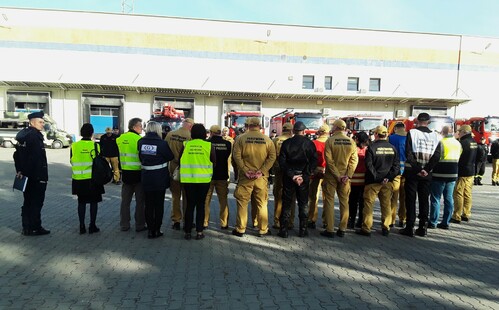 Inspektor Wojewódzkiego Inspektoratu Ochrony Środowiska w Warszawie oraz kilkunastu przedstawicieli innych służb stoi na placu. W tle wozy straży pożarnej stojące na tle jednopiętrowej hali.