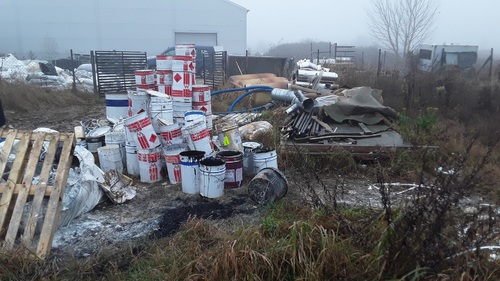 Białe, metalowe pojemniki złożone w nieładzie, jedne na drugich, na otwartym terenie. W tle znajduje się hala.